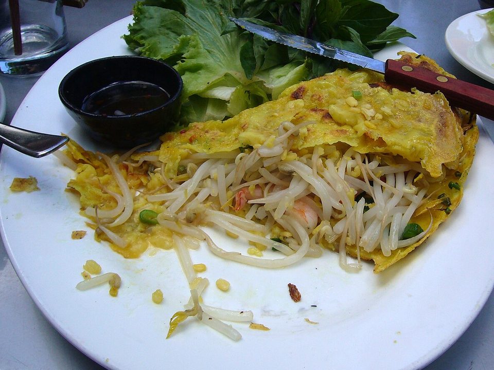 Goldgelbe Bánh-xèo-Crêpe mit Sprossen auf einem Teller, dahinter Salat und Dip