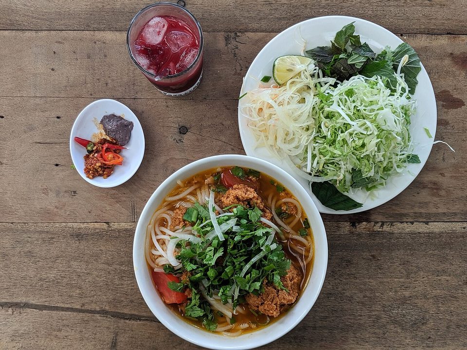 Schale Bún riêu mit roter Brühe und Kräutern, daneben ein Teller mit Gemüse