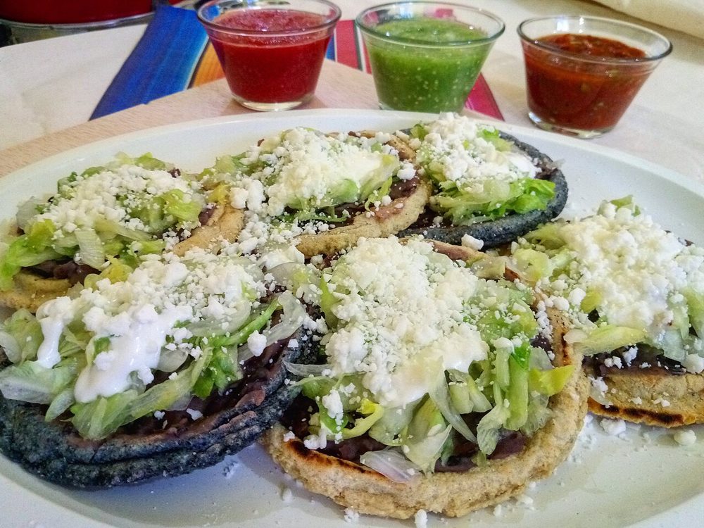 Mehrere Sopes mit Bohnen, Salat und Käse, dahinter drei Salsas